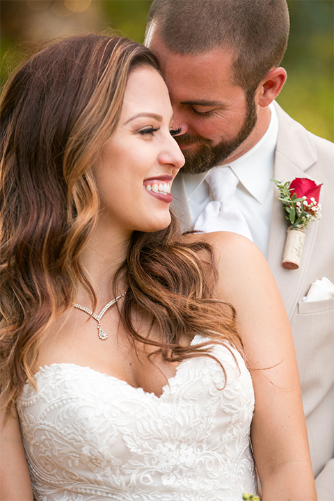 Temecula outdoor rustic wedding at lake oak meadows bride form fitting strapless gown with sweetheart neckline and lace detail with long veil and groom tan notch lapel suit with matching vest and white dress shirt and long white tie with red floral boutonniere hugging and close up bride holding white and red floral bridal bouquet with red ribbon decor