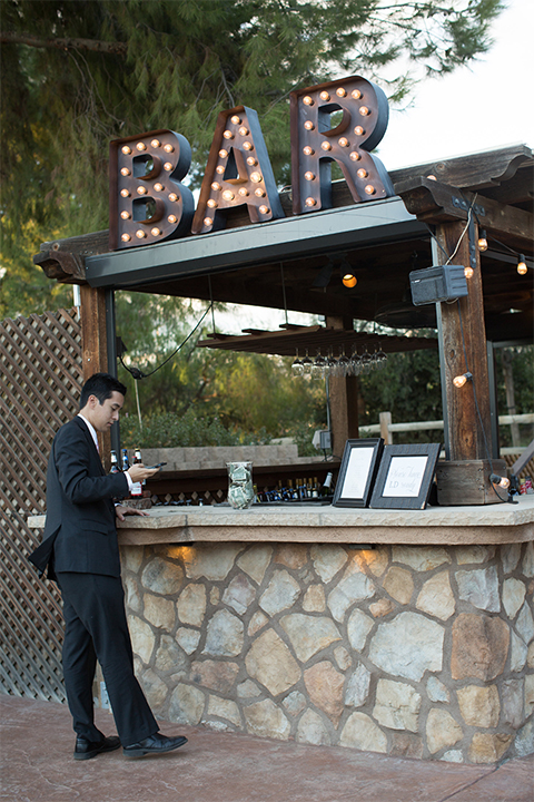 Temecula outdoor rustic wedding at the lake oak meadows table set up with white table linen and white and red flower decor with greenery florals and white initial letters with white place settings and gold table numbers with bar