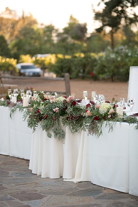 Temecula outdoor rustic wedding at the lake oak meadows table set up with white table linen and white and red flower decor with greenery florals and white initial letters with white place settings and gold table numbers