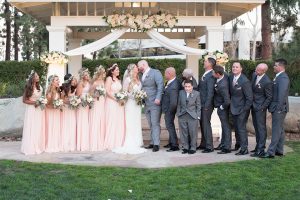 Orange county outdoor wedding shoot at turnip rose garden bride form fitting gown with lace detail and long sleeves with long veil and sweetheart neckline with groom heather grey notch lapel suit with matching vest and white dress shirt with long white tie and blush pink floral boutonniere kissing with wedding party bridesmaids long blush pink dresses with flower crowns and groomsmen charcoal grey suits with long blush pink ties