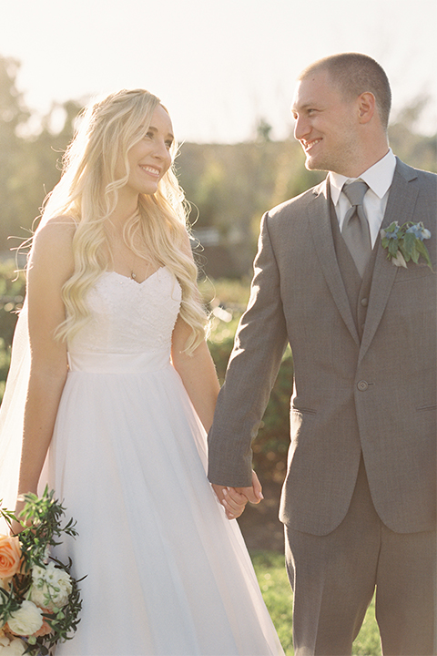 Southern california wedding at riverview free church bride strapless ball gown with a sweetheart neckline and tulle skirt with a long veil and braided hairstyle with groom grey notch lapel suit with matching vest and white dress shirt with long grey tie and light blue and green floral boutonniere holding hands