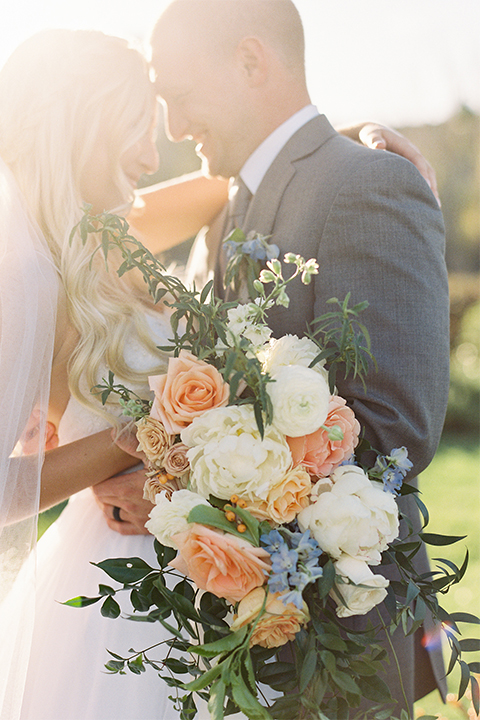 Southern california wedding at riverview free church bride strapless ball gown with a sweetheart neckline and tulle skirt with a long veil and braided hairstyle with groom grey notch lapel suit with matching vest and white dress shirt with long grey tie and light blue and green floral boutonniere hugging and bride holding white and orange floral bridal bouquet