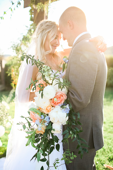 Southern california wedding at riverview free church bride strapless ball gown with a sweetheart neckline and tulle skirt with a long veil and braided hairstyle with groom grey notch lapel suit with matching vest and white dress shirt with long grey tie and light blue and green floral boutonniere hugging and bride holding white and orange floral bridal bouquet