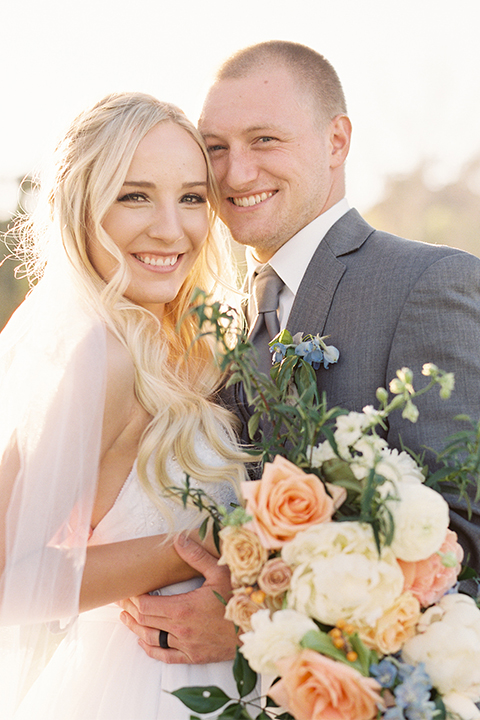 Southern california wedding at riverview free church bride strapless ball gown with a sweetheart neckline and tulle skirt with a long veil and braided hairstyle with groom grey notch lapel suit with matching vest and white dress shirt with long grey tie and light blue and green floral boutonniere hugging and bride holding white and orange floral bridal bouquet