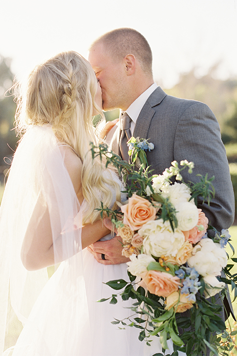 Southern california wedding at riverview free church bride strapless ball gown with a sweetheart neckline and tulle skirt with a long veil and braided hairstyle with groom grey notch lapel suit with matching vest and white dress shirt with long grey tie and light blue and green floral boutonniere hugging and kissing and bride holding white and orange floral bridal bouquet