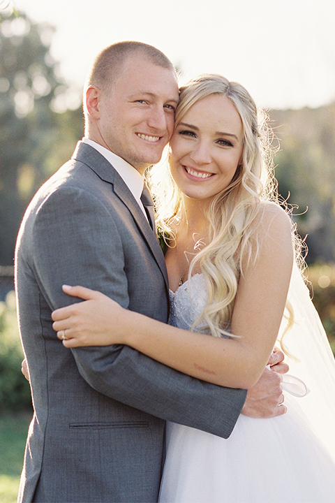 Southern california wedding at riverview free church bride strapless ball gown with a sweetheart neckline and tulle skirt with a long veil and braided hairstyle with groom grey notch lapel suit with matching vest and white dress shirt with long grey tie and light blue and green floral boutonniere hugging