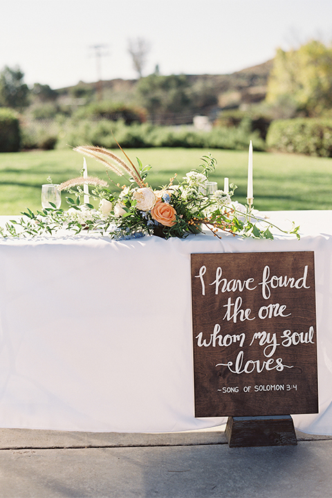 Southern california outdoor wedding at the riverview free church wedding welcome sign light brown wood sign with white calligraphy writing rustic decor with white and green flowers on top with table white linen and white place settings with tall white candles and gold holders with light orange and white flower centerpiece decor with gold vases and gold silverware
