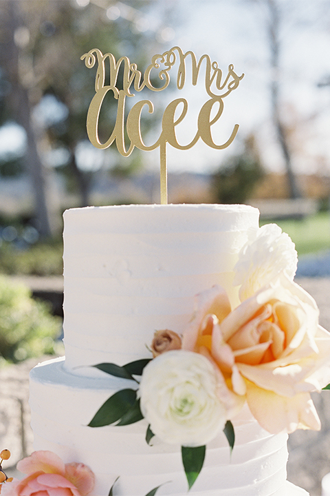 Southern california outdoor wedding at the riverview free church wedding welcome sign light brown wood sign with white calligraphy writing rustic decor with white and green flowers on top with table white linen and white place settings with tall white candles and gold holders with light orange and white flower centerpiece decor with gold vases and gold silverware with three tier white wedding cake with flower decor and gold calligraphy cake topper