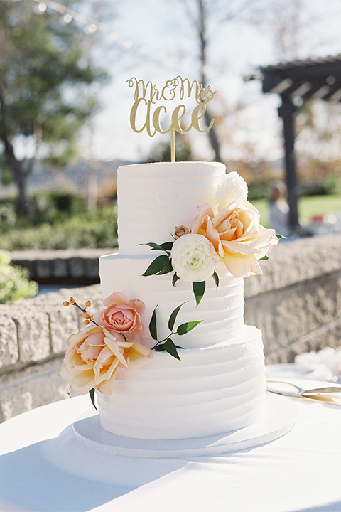 Southern california outdoor wedding at the riverview free church wedding welcome sign light brown wood sign with white calligraphy writing rustic decor with white and green flowers on top with table white linen and white place settings with tall white candles and gold holders with light orange and white flower centerpiece decor with gold vases and gold silverware with three tier white wedding cake with flower decor and gold calligraphy cake topper