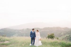 Santa barbara citrus inspired wedding shoot bride a line ball gown with lace detail on the bodice and a high neckline with long sleeves and groom cobalt blue notch lapel suit with matching vest and white dress shirt with long black skinny tie and white floral boutonniere walking and holding hands bride holding yellow and orange floral bridal bouquet outside