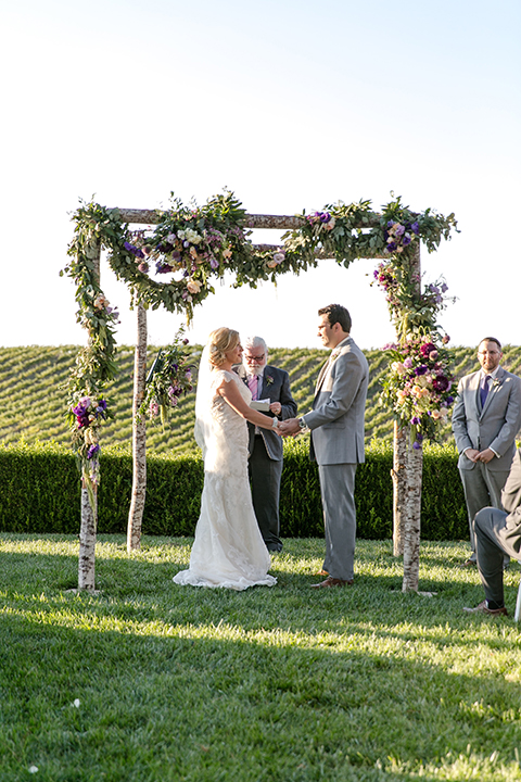Temecula outdoor wedding at callaway winery bride form fitting lace gown with sheer back design and high neckline with groom heather grey notch lapel suit with matching vest and white dress shirt with long white tie and light purple floral boutonniere holding hands during ceremony
