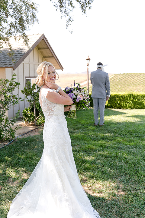 Temecula outdoor wedding at callaway winery bride form fitting lace gown with sheer back design and high neckline with groom heather grey notch lapel suit with matching vest and white dress shirt with long white tie and light purple floral boutonniere first look