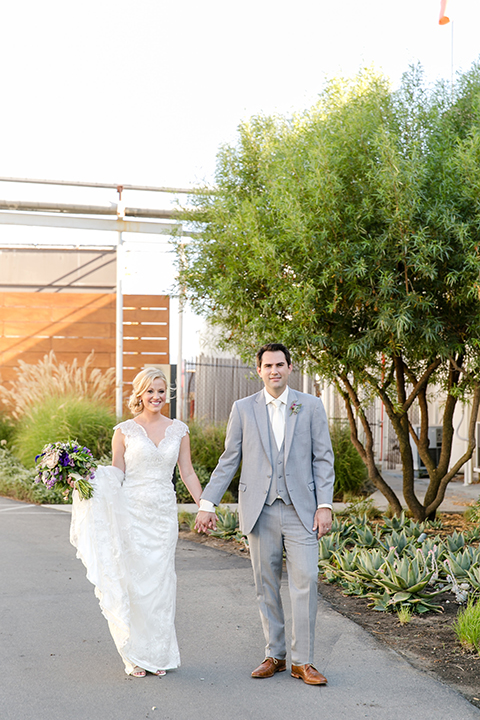 Temecula outdoor wedding at callaway winery bride form fitting lace gown with sheer back design and high neckline with groom heather grey notch lapel suit with matching vest and white dress shirt with long white tie and light purple floral boutonniere holding hands