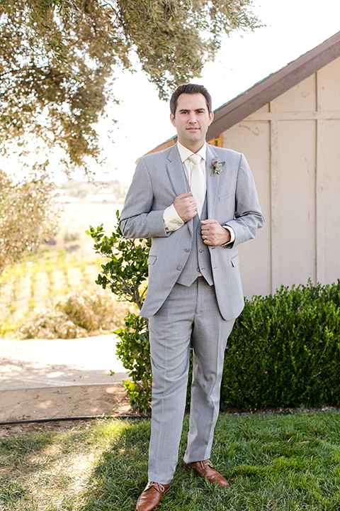 Temecula outdoor wedding at callaway winery groom heather grey notch lapel suit with a matching vest and white dress shirt with a long white tie and light purple floral boutonniere standing holding jacket