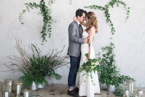 Los angeles minimal and classic wedding shoot at fd studio bride a line strapless gown with straight neckline and lace skirt with pockets and simple design with groom grey notch lapel suit with white dress shirt and long black skinny tie with a white floral boutonniere standing and hugging during ceremony bride holding green floral bridal bouquet