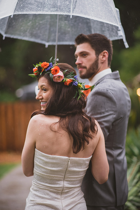 Temecula outdoor wedding at temecula creek inn bride form fitting simple strapless gown with a straight neckline and teal rain boots with groom grey notch lapel suit with a white dress shirt and long brown tie with an orange floral boutonniere hugging and holding clear umbrella