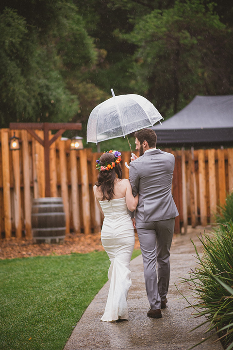 Temecula outdoor wedding at temecula creek inn bride form fitting simple strapless gown with a straight neckline and teal rain boots with groom grey notch lapel suit with a white dress shirt and long brown tie with an orange floral boutonniere holding hands and walking under clear umbrella