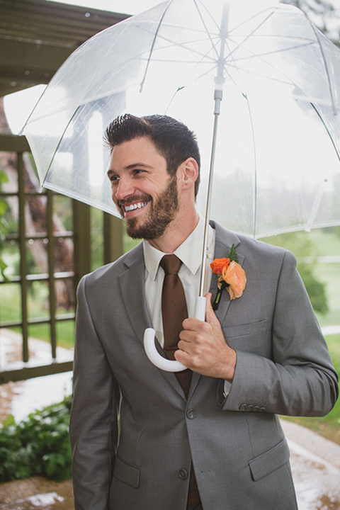 Temecula outdoor wedding at temecula creek inn groom grey notch lapel suit with a white dress shirt and long brown tie with yellow and orange floral boutonniere holding clear umbrella