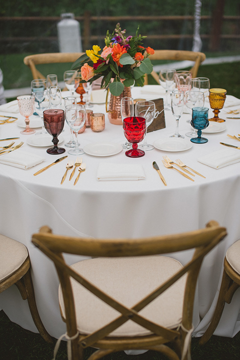 Temecula outdoor wedding at temecula creek inn table set up light brown wood table with matching wood chairs and colorful flower centerpiece decor with colorful glassware and white place settings with brown wood table numbers with white calligraphy writing