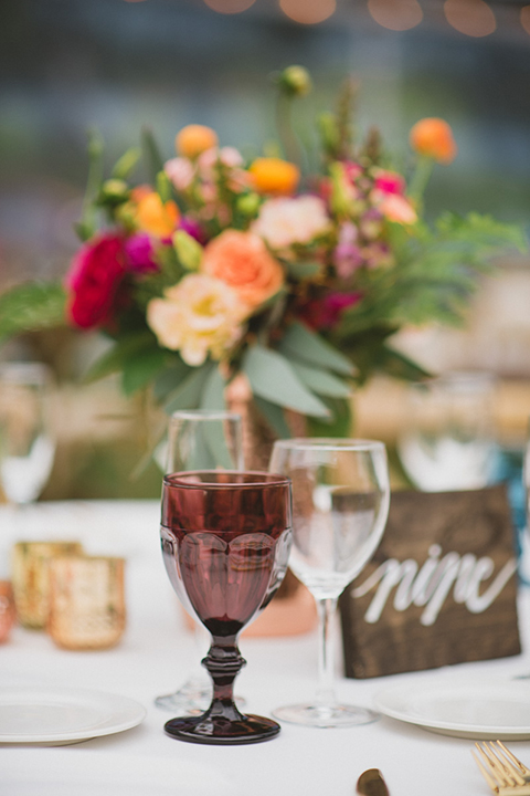 Temecula outdoor wedding at temecula creek inn table set up light brown wood table with matching wood chairs and colorful flower centerpiece decor with colorful glassware and white place settings with brown wood table numbers with white calligraphy writing
