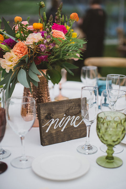 Temecula outdoor wedding at temecula creek inn table set up light brown wood table with matching wood chairs and colorful flower centerpiece decor with colorful glassware and white place settings with brown wood table numbers with white calligraphy writing