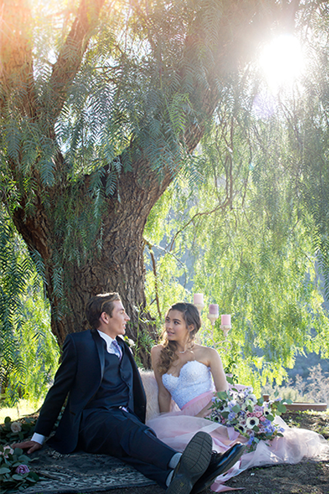 San diego outdoor woodland wedding shoot bride blush pink chiffon strapless gown with a sweetheart neckline and groom navy blue shawl lapel tuxedo with a matching vest and white dress shirt with a long pink striped tie and white floral boutonniere sitting on blanket under tree
