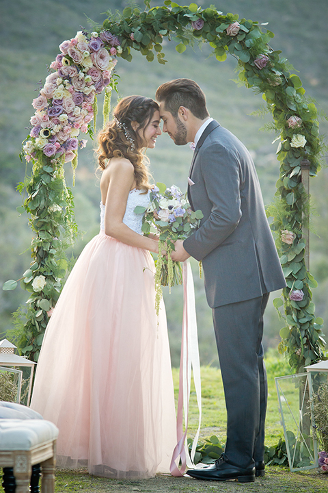 San diego outdoor woodland wedding shoot bride blush pink chiffon strapless gown with a sweetheart neckline and groom navy blue shawl lapel tuxedo with a matching vest and white dress shirt with a long pink striped tie and white floral boutonniere holding hands during ceremony and bride holding white floral bridal bouquet