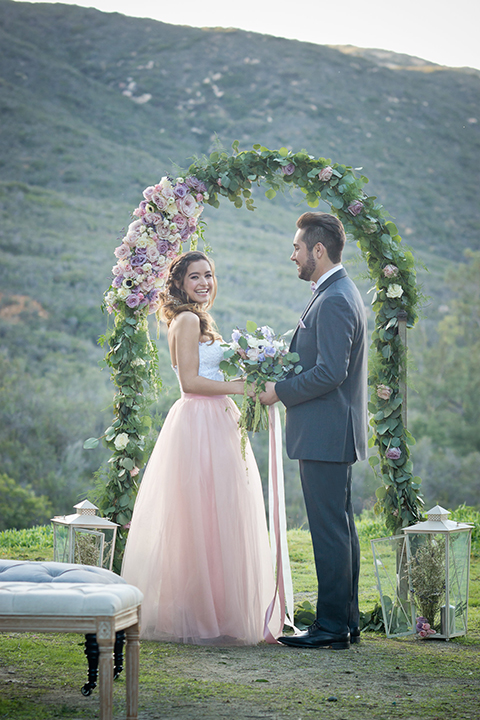 San diego outdoor woodland wedding shoot bride blush pink chiffon strapless gown with a sweetheart neckline and groom navy blue shawl lapel tuxedo with a matching vest and white dress shirt with a long pink striped tie and white floral boutonniere holding hands during ceremony and bride holding white floral bridal bouquet