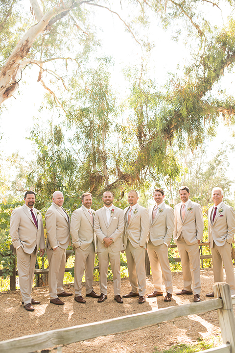 Temecula outdoor rustic wedding at lake oak meadows groom tan notch lapel suit with matching vest and white dress shirt and long white tie with red floral boutonniere standing with groomsmen tan suits