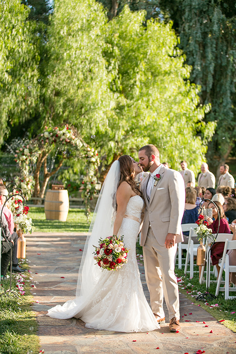 Temecula outdoor rustic wedding at lake oak meadows bride form fitting strapless gown with sweetheart neckline and lace detail with long veil and groom tan notch lapel suit with matching vest and white dress shirt and long white tie with red floral boutonniere hugging and close up bride holding white and red floral bridal bouquet with red ribbon decor kissing after ceremony
