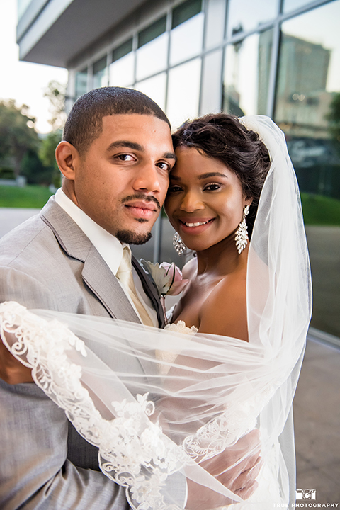 San diego outdoor wedding at ultimate skybox bride form fitting strapless lace gown with a sweetheart neckline and crystal belt with long veil and groom heather grey notch lapel suit with a white dress shirt and matching vest with a long white tie and white floral boutonniere hugging