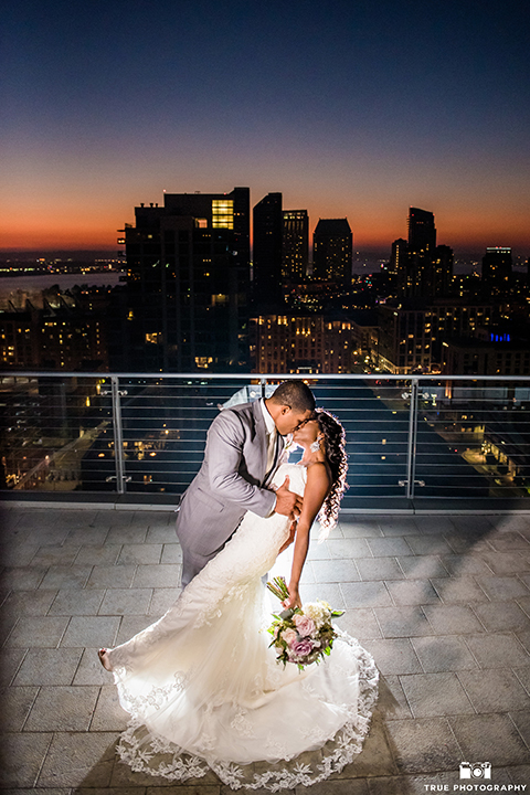 San diego outdoor wedding at ultimate skybox bride form fitting strapless lace gown with a sweetheart neckline and crystal belt with long veil and groom heather grey notch lapel suit with a white dress shirt and matching vest with a long white tie and white floral boutonniere standing and kissing at night bride holding white floral bridal bouquet