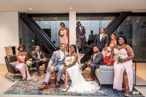 San diego outdoor wedding at ultimate skybox bride form fitting strapless lace gown with a sweetheart neckline and crystal belt with long veil and groom heather grey notch lapel suit with a white dress shirt and matching vest with a long white tie and white floral boutonniere sitting with wedding party bridesmaids long blush pink dresses and groomsmen charcoal grey suits with long white ties