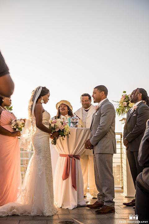 San diego outdoor wedding at ultimate skybox bride form fitting strapless lace gown with a sweetheart neckline and crystal belt with long veil and groom heather grey notch lapel suit with a white dress shirt and matching vest with a long white tie and white floral boutonniere standing during ceremony bride holding white floral bridal bouquet
