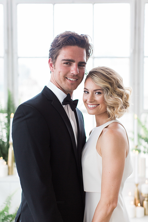 Downtown los angeles wedding shoot at fd photo studio bride form fitting simple gown with a high halter neckline and groom black notch lapel tuxedo by michael kors with a white dress shirt and black bow tie smiling and holding hands