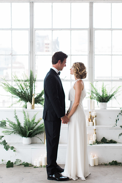 Downtown los angeles wedding shoot at fd photo studio bride form fitting simple gown with a high halter neckline and groom black notch lapel tuxedo by michael kors with a white dress shirt and black bow tie holding hands with greenery decor
