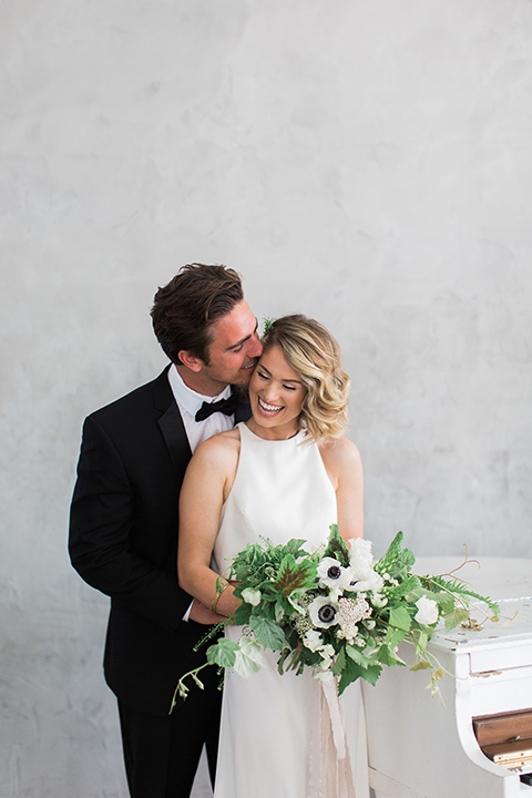 Downtown los angeles wedding shoot at fd photo studio bride form fitting simple gown with a high halter neckline and groom black notch lapel tuxedo by michael kors with a white dress shirt and black bow tie hugging and smiling bride holding white and green floral bridal bouquet
