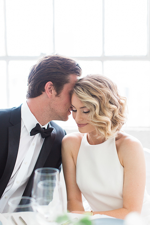 Downtown los angeles wedding shoot at fd photo studio bride form fitting simple gown with a high halter neckline and groom black notch lapel tuxedo by michael kors with a white dress shirt and black bow tie sitting at table hugging and smiling