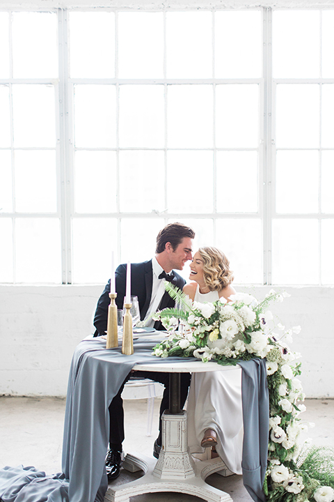 Downtown los angeles wedding shoot at fd photo studio bride form fitting simple gown with a high halter neckline and groom black notch lapel tuxedo by michael kors with a white dress shirt and black bow tie sitting at table smiling