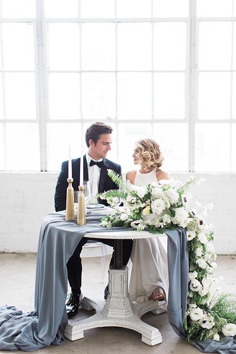 Downtown los angeles wedding shoot at fd photo studio bride form fitting simple gown with a high halter neckline and groom black notch lapel tuxedo by michael kors with a white dress shirt and black bow tie sitting at table smiling