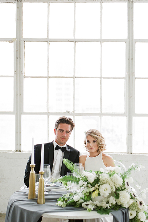 Downtown los angeles wedding shoot at fd photo studio bride form fitting simple gown with a high halter neckline and groom black notch lapel tuxedo by michael kors with a white dress shirt and black bow tie sitting at table