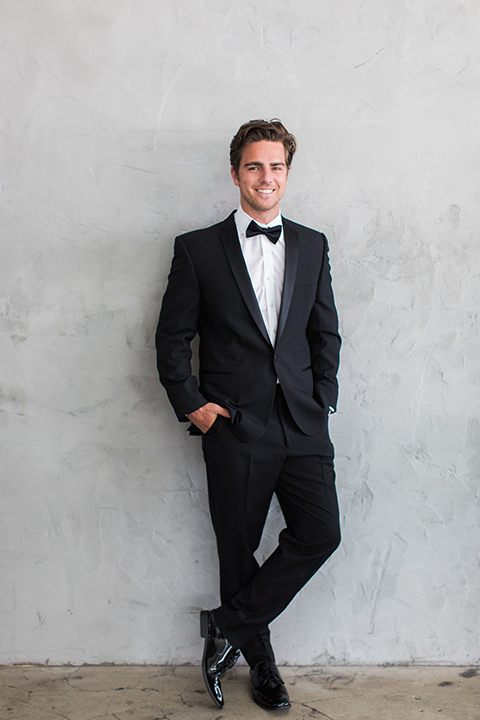 Downtown los angeles wedding shoot at fd photo studio groom black notch lapel tuxedo by michael kors with a white dress shirt and black bow tie standing against wall with hands in pockets