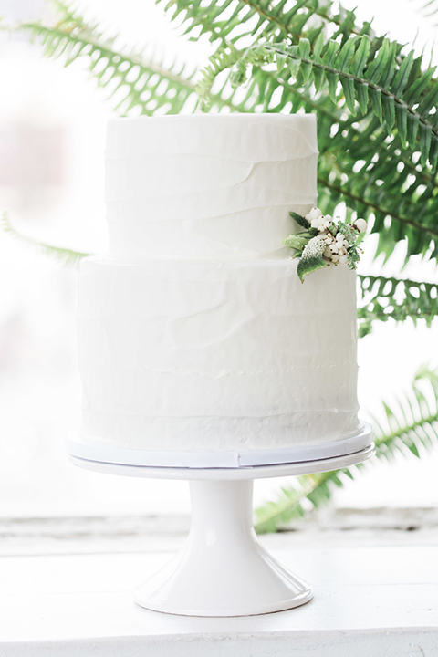 Downtown los angeles wedding shoot at fd photo studio table set up with grey table linen and white and green flower decor with clear ghost chairs and white and grey place settings with white candle decor and piano with two tier white wedding cake with white and green flower decor