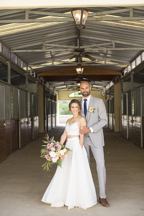 Los angeles outdoor wedding at brookview ranch bride two piece wedding dress with tulle skirt and crop top bodice with thin straps and a sweetheart neckline and groom heather grey notch lapel suit with a matching vest and white dress shirt with a long navy blue tie and white floral boutonniere hugging and holding hands