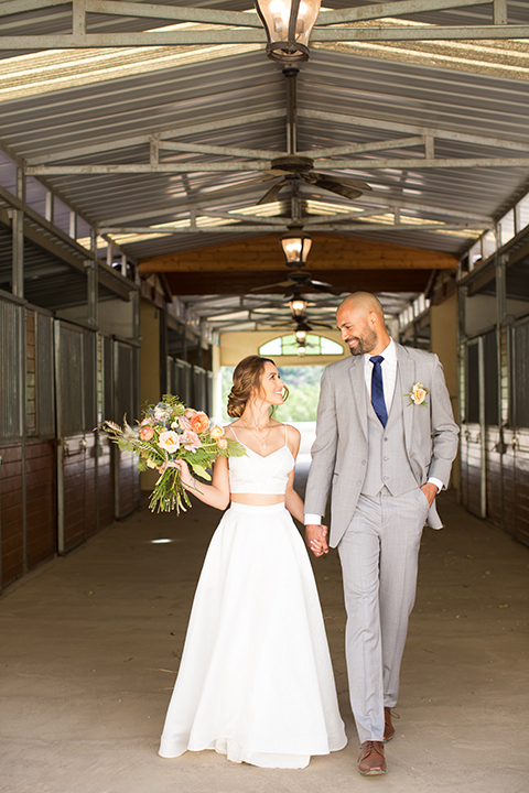 Los angeles outdoor wedding at brookview ranch bride two piece wedding dress with tulle skirt and crop top bodice with thin straps and a sweetheart neckline and groom heather grey notch lapel suit with a matching vest and white dress shirt with a long navy blue tie and white floral boutonniere holding hands and bride holding white and pink floral bridal bouquet