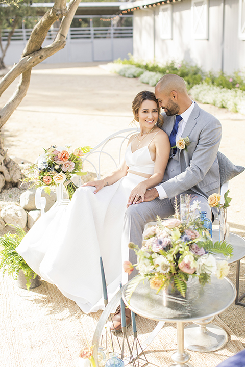 Los angeles outdoor wedding at brookview ranch bride two piece wedding dress with tulle skirt and crop top bodice with thin straps and a sweetheart neckline and groom heather grey notch lapel suit with a matching vest and white dress shirt with a long navy blue tie and white floral boutonniere sitting on white couch with white and pink flower decor