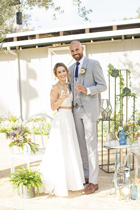 Los angeles outdoor wedding at brookview ranch bride two piece wedding dress with tulle skirt and crop top bodice with thin straps and a sweetheart neckline and groom heather grey notch lapel suit with a matching vest and white dress shirt with a long navy blue tie and white floral boutonniere standing and holding drinks by wedding table and decor