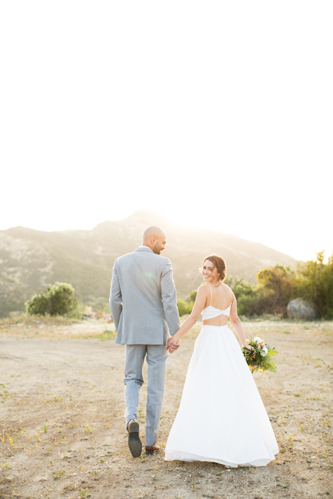 Los angeles outdoor wedding at brookview ranch bride two piece wedding dress with tulle skirt and crop top bodice with thin straps and a sweetheart neckline and groom heather grey notch lapel suit with a matching vest and white dress shirt with a long navy blue tie and white floral boutonniere holding hands and bride holding white and pink floral bridal bouquet