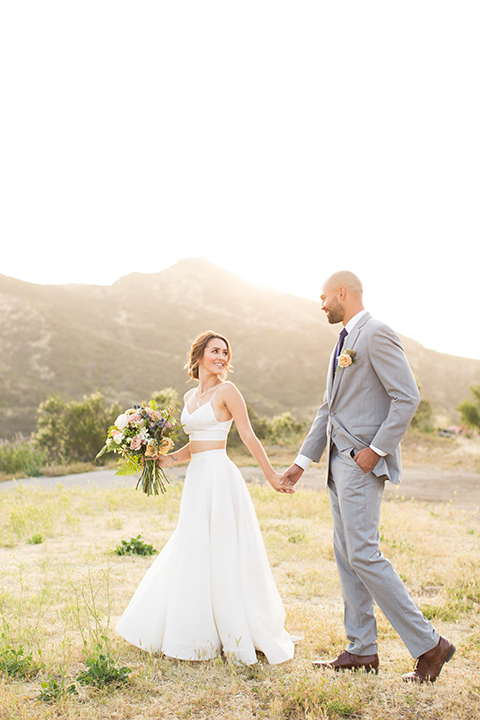 Los angeles outdoor wedding at brookview ranch bride two piece wedding dress with tulle skirt and crop top bodice with thin straps and a sweetheart neckline and groom heather grey notch lapel suit with a matching vest and white dress shirt with a long navy blue tie and white floral boutonniere holding hands and bride holding white and pink floral bridal bouquet