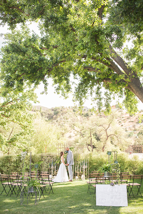 Los angeles outdoor wedding at brookview ranch bride two piece wedding dress with tulle skirt and crop top bodice with thin straps and a sweetheart neckline and groom heather grey notch lapel suit with a matching vest and white dress shirt with a long navy blue tie and white floral boutonniere holding hands during ceremony with backdrop and flower decor
